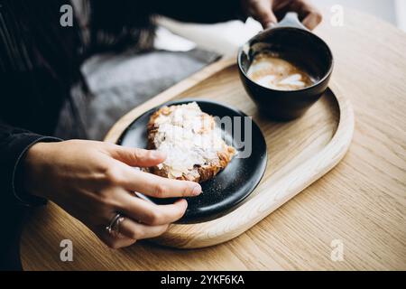 Cropped view of an unrecognizable person's hands holding a delicious almond croissant and a cup of coffee, served on a wooden table in a cozy cafe set Stock Photo