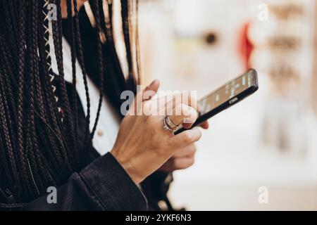 A stylish cropped unrecognizable caucasian woman with braids is captured using her smartphone in a cafe. The focus is on her hands holding the device, Stock Photo