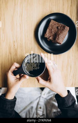 Top view of a cropped unrecognizable woman's hands holding a black coffee cup, with a chocolate brownie on a black plate beside her on a wooden table. Stock Photo