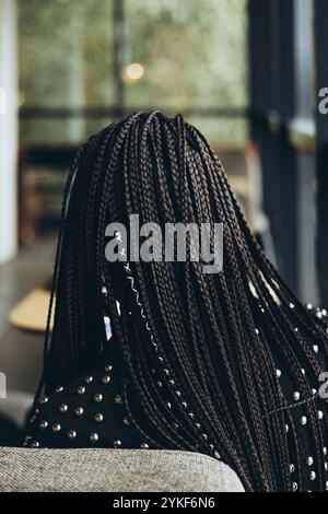 Back view of an unrecognizable stylish Caucasian woman wearing a studded jacket, showing detailed braided hair, sitting in a cafe with a serene atmosp Stock Photo