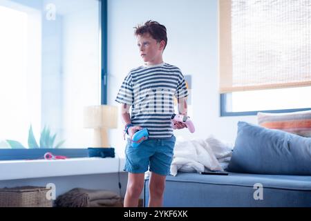 A preteen boy stands in a bright living room holding a game controller in each hand He looks to the side, thinking about his next game Stock Photo