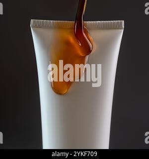 Close-up view of a white cosmetic tube with a gooey, caramel like substance oozing over the edge against a dark background. Stock Photo