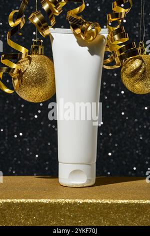 A white cosmetic tube stands surrounded by shimmering golden Christmas ornaments and ribbons, against a sparkling black background, embodying a luxuri Stock Photo