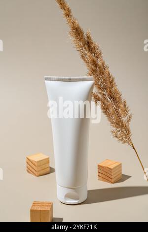 A blank white plastic hand cream tube stands on a beige background, flanked by ornamental dried grass and wooden blocks, perfect for branding presenta Stock Photo