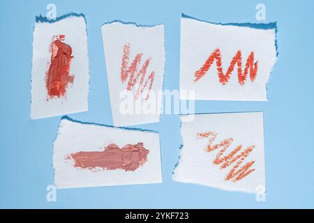 Various shades of lipstick are swatched on torn paper pieces, set against a pastel blue background The image displays rich textures and vibrant colors Stock Photo