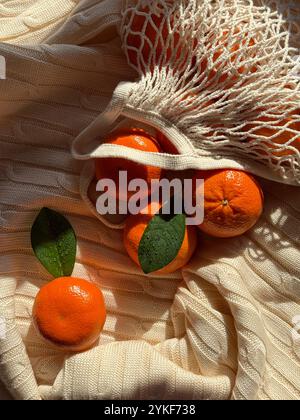 Top view of fresh, juicy mandarins with green leaves in a white mesh bag, placed on a textured cream fabric, illuminated by warm sunlight creating a c Stock Photo