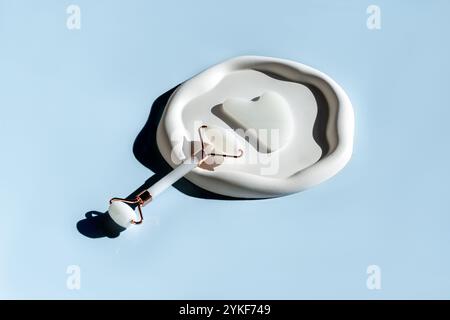 From above, a white face roller and a Gua Sha massage tool lying on an elegant marble dish, set against a soothing blue background. These skincare too Stock Photo