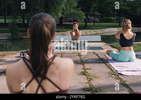 Outdoor yoga session with a woman instructor demonstrating the Lotus pose or Padmasana to students in a park. Multiple participants, including two vis Stock Photo