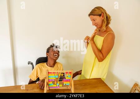 A joyful young Black boy with cerebral palsy engages with colorful abacus beads during a playful learning session with his Caucasian female teacher at Stock Photo