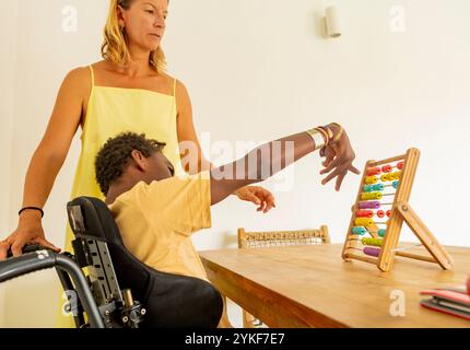 A young African American boy with cerebral palsy engages with a colorful abacus under the guidance of his teacher at a holiday home. The boy, seated i Stock Photo