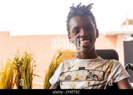 A joyful young African American boy with cerebral palsy, sitting in his wheelchair, smiles brightly while looking away. Surrounded by lush green plant Stock Photo