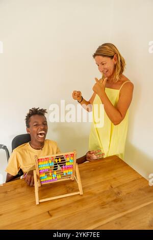 A joyful young Black boy with cerebral palsy engages with colorful abacus beads during a playful learning session with his Caucasian female teacher at Stock Photo