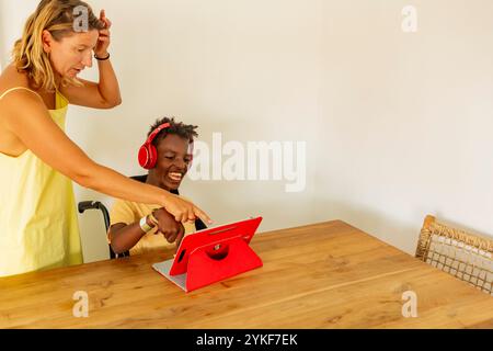 A young Black boy with cerebral palsy interacts joyfully with his female teacher at a holiday home. He uses a tablet and wears headphones while she as Stock Photo