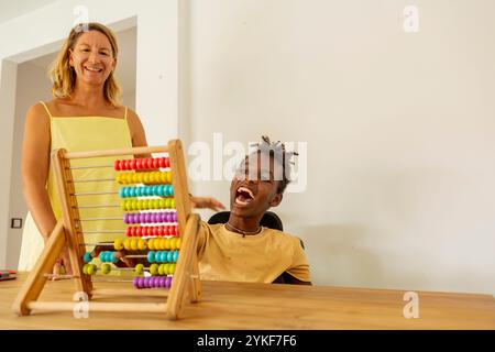 A joyful young Black boy with cerebral palsy engages with colorful abacus beads during a playful learning session with his Caucasian female teacher at Stock Photo