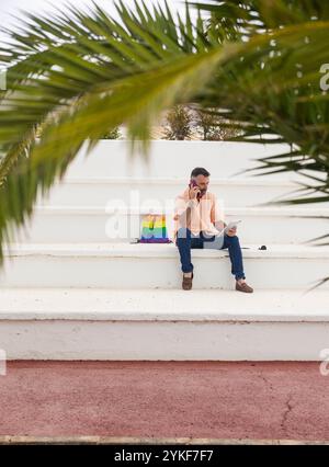 Adult male sitting on white steps, talking on the phone and looking at a tablet, with a rainbow-colored bag beside him, under palm fronds Stock Photo