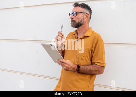 A contemplative gay man with a beard, wearing glasses and a pride bracelet, stands holding a tablet and digital pen against a white wall Stock Photo