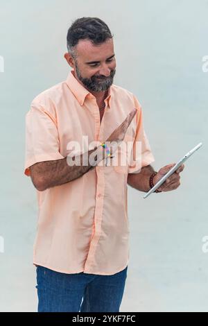 A mature bearded man in an orange shirt smiles while interacting with a tablet, wearing a rainbow bracelet symbolizing LGBTQIA+ support Stock Photo