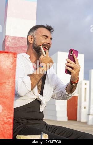 Cheerful and expressive transvestite gay man using his smartphone outdoors While applying lipstick, she wears a vibrant outfit that reflects his LGBTQ Stock Photo