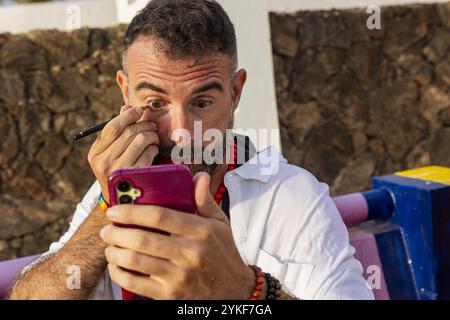 A transvestite gay man concentrates on applying eyeliner using his smartphone as a mirror in an outdoor setting, showcasing individuality and expressi Stock Photo
