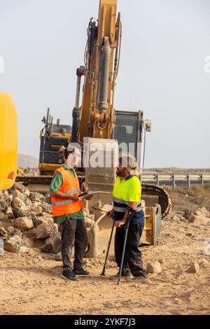 A disabled inspector on crutches and his colleague, both wearing reflective vests, talk about safety near an excavator at a dusty renewable energy con Stock Photo