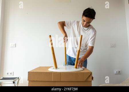 A young trans man is engaged in assembling a wooden table in his bright, new apartment, symbolizing a fresh start and self-reliance Stock Photo