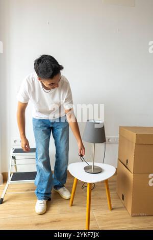 A trans man arranges a lamp on a side table in his new apartment, surrounded by moving boxes, reflecting independence and a new beginning Stock Photo