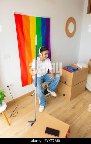 Trans man surrounded by moving boxes in his new home, holding a broom and listening to music through headphones A rainbow pride flag hangs on the wall Stock Photo