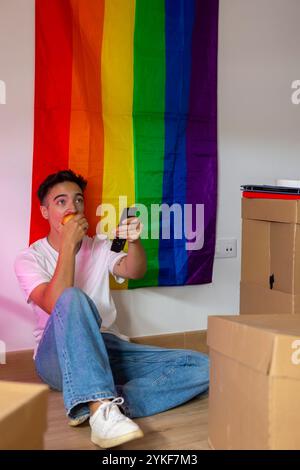 A trans man takes a break from his movement, sitting in a room adorned with a pride flag, holding a remote control, surrounded by cardboard boxes and Stock Photo