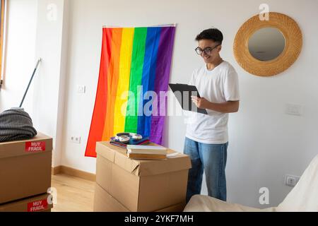 Cheerful trans man smiles while reading a document, surrounded by moving boxes in his new apartment, a vibrant pride flag hanging on the wall Stock Photo