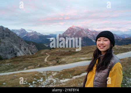 A joyful asian tourist stands against the breathtaking backdrop of the Dolomites in Italy, with rugged mountains and a colorful sky creating a serene Stock Photo