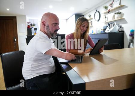 A disability man collaborates with a woman at a home They are both working on laptops at a wooden table in a modern kitchen, demonstrating teamwork an Stock Photo