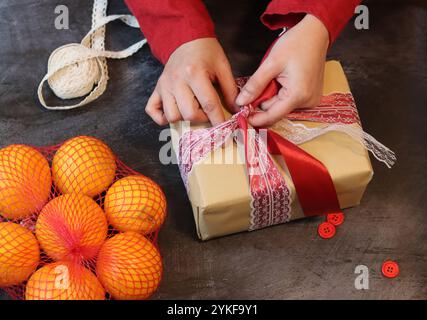 Hands of a young woman wearing red dress wrapping Christmas gifts on a wooden background. Gift box with red ribbon and tangerines top view photo Stock Photo
