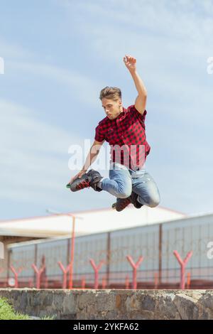 A young man is captured mid-air performing an impressive jump while rollerblading on inline skates against a clear blue sky and industrial backdrop. Stock Photo
