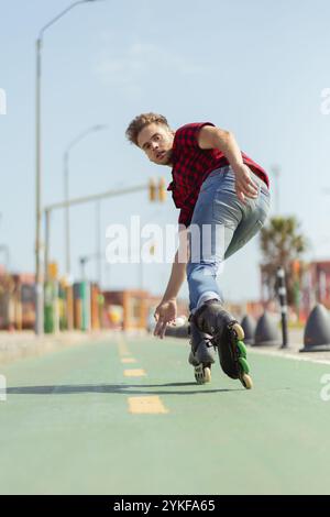A young man engages in inline skating on a city bike lane, performing with agility and skill under clear blue skies. He looks towards the camera, exud Stock Photo
