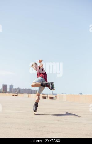 A young man shows off his skills by performing a jumping stunt on inline skates at an outdoor skate park. He wears a red plaid shirt and shorts, and l Stock Photo