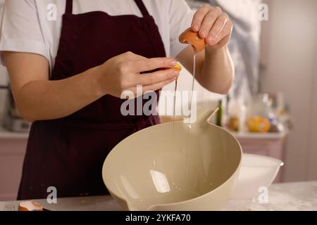 An intimate scene in a kitchen where a person in a maroon apron cracks an egg into a bowls The focus is on the hands and the egg, set against a softly Stock Photo