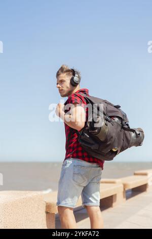 A thoughtful young man with a backpack and headphones is casually walking by the seaside, looking away from the camera, under a clear blue sky. Stock Photo