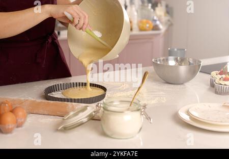 A person in a maroon apron pours batter into a tart pan in a well-equipped kitchen, suggesting a cozy, homemade baking scene Stock Photo