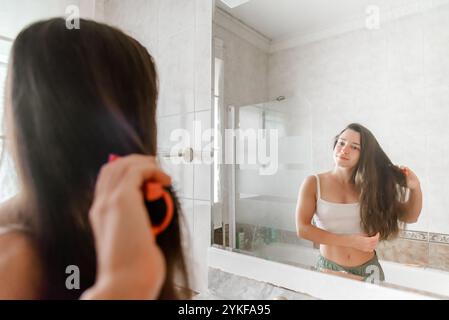 A young woman gently combs her long hair while looking at her reflection in a bright, tiled bathroom mirror. Stock Photo
