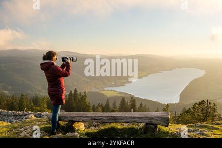 Back view of unrecognizable woman using a camera to capture the serene sunset in Dent de Vaulion, overlooking a scenic Swiss lake. Stock Photo