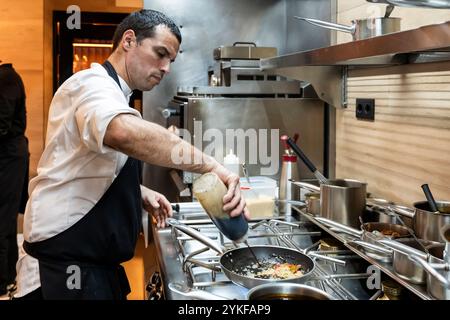 A focused chef in a restaurant kitchen is intently cooking and seasoning a dish Equipment and ingredients surround him in this dynamic restaurant envi Stock Photo