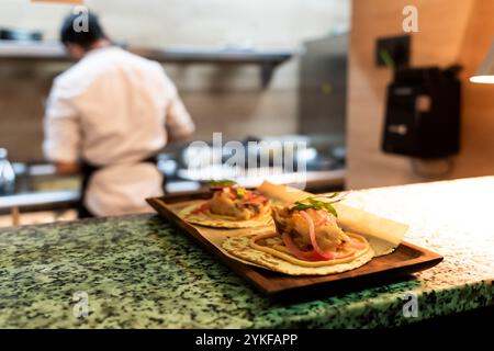 Two freshly prepared tacos sit on a wooden tray, ready to be served, as a chef works in the kitchen in the background The scene highlights the artistr Stock Photo