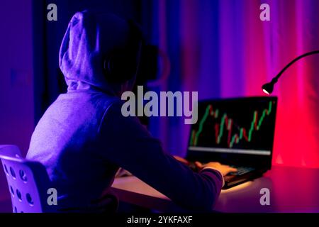 Back view of unrecognizable boy trading stocks on a laptop, highlighted by colorful screen light in a dark room. The image captures a moment of concen Stock Photo