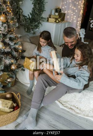 A father and two daughters share the joy of Christmas morning, opening presents on a bed. The festive atmosphere is filled with warmth and laughter Stock Photo