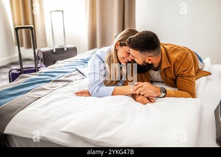Young happy couple is on vacation. Tourists arrived in hotel room and relaxing on bed. Stock Photo