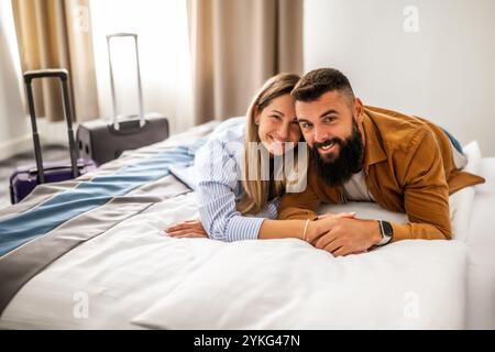 Young happy couple is on vacation. Tourists arrived in hotel room and relaxing on bed. Stock Photo