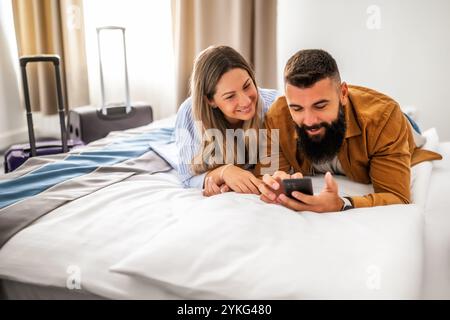 Young happy couple is on vacation. Tourists arrived in hotel room with their suitcases. They are lying on bed and using smartphone. Stock Photo
