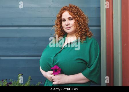 Plus Size Woman with Curly Red Hair in Green Clothing Holds Flower Against Vibrant Blue Wall Background, Beauty and Confidence Outdoors Stock Photo
