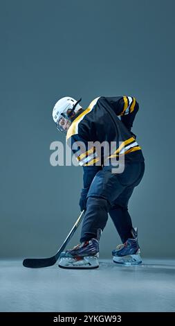 Vertical full-length image of man, hockey player in helmet and dark uniform, standing with stick, ready to hit the puck. Side view Stock Photo