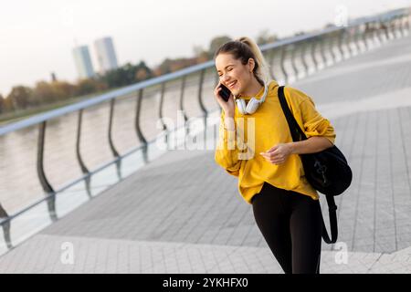 A cheerful woman dressed in a vibrant yellow sweater laughs on her phone while strolling along a picturesque riverside walkway, enjoying the warm suns Stock Photo